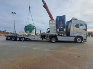 Camión con plataforma cargando equipo cubierto, usando grúa para levantar carga