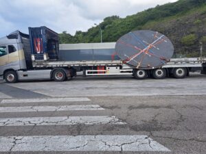 Camión transportando gran disco metálico asegurado con correas en plataforma.