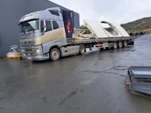 Camión transportando grandes piezas metálicas curvas sobre plataforma en zona industrial